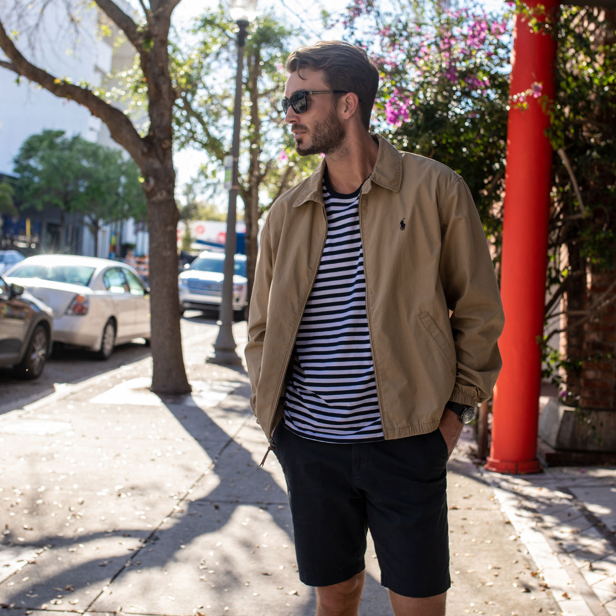 A man is stood on a beach in Miami. He's wearing a thin jacket, a striped t-shirt and black shorts.