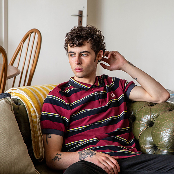 A man is sat on a green couch in a living room. He is wearing a red and blue striped polo shirt. 