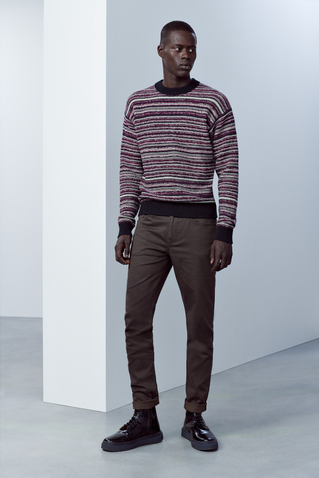 A man standing next to a white washed wall. He is wearing a stripey jumper from Hugo and some dark Hugo jeans, matched with black Hugo brogues. 